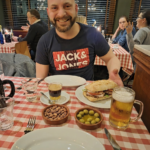 Noe Serrano, pintor de coches emigrado a Alemania, sonriendo en un restaurante ante un auténtico almuerzo valenciano con bocadillo Chivito, cremaet, cacaos y olivas, celebrando sus primeros resultados con Partner 360, en el futuro.