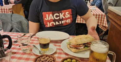 Noe Serrano, pintor de coches emigrado a Alemania, sonriendo en un restaurante ante un auténtico almuerzo valenciano con bocadillo Chivito, cremaet, cacaos y olivas, celebrando sus primeros resultados con Partner 360, en el futuro.