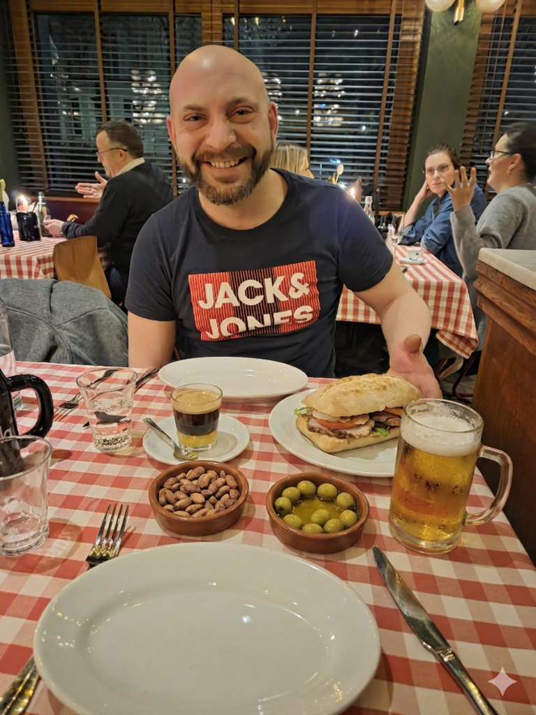 Noe Serrano, pintor de coches emigrado a Alemania, sonriendo en un restaurante ante un auténtico almuerzo valenciano con bocadillo Chivito, cremaet, cacaos y olivas, celebrando sus primeros resultados con Partner 360, en el futuro.