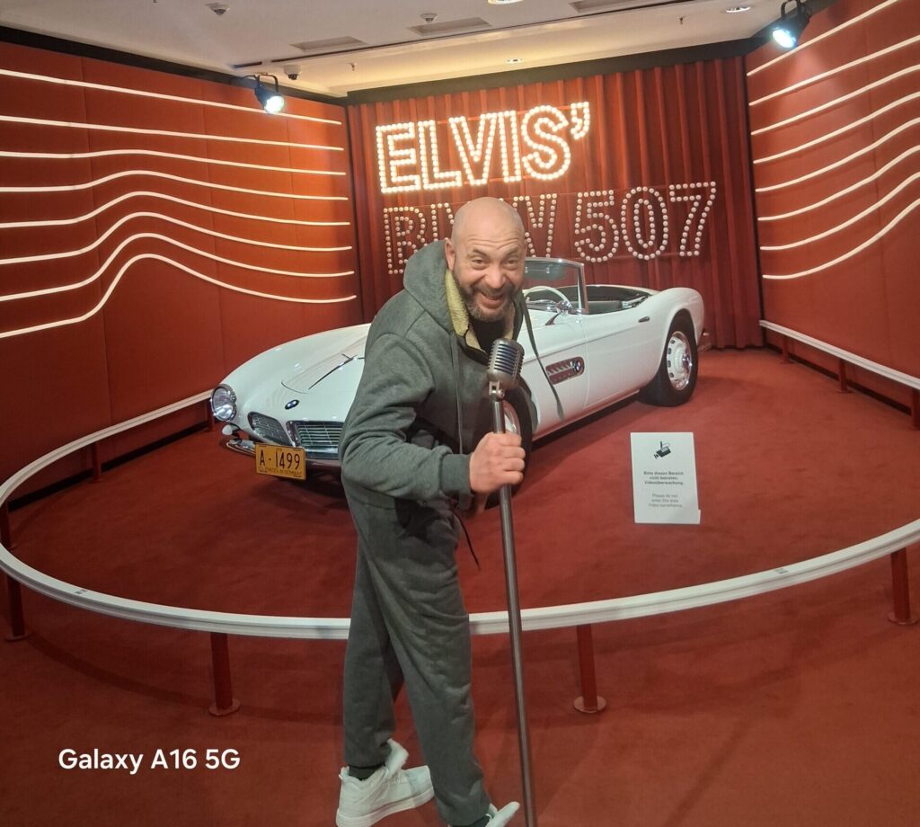 Noé Serrano cantando con un micrófono de Elvis Presley en su BMW 507 blanco en el Museo BMW de Múnich, ilustrando el inicio de la Ruta 2032.