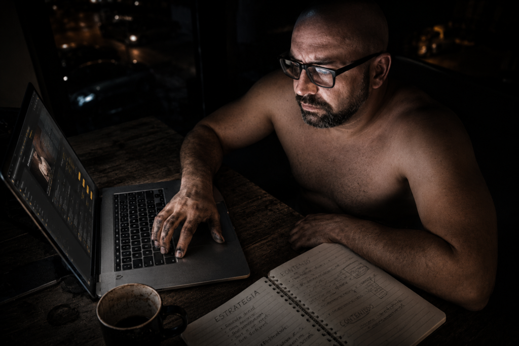 Hombre con gafas trabajando de noche frente a un portátil con las manos manchadas de grasa de taller, analizando cuánto se tarda en ganar dinero online en su libreta de estrategia.