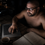 Hombre con gafas trabajando de noche frente a un portátil con las manos manchadas de grasa de taller, analizando cuánto se tarda en ganar dinero online en su libreta de estrategia.