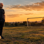 Noé Serrano de pie y solo en un campo de fútbol modesto al atardecer, con un balón a los pies, representando la metáfora de la Intertoto sobre mantener la constancia sin ver resultados